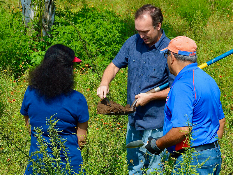 A man in a field holding a shovel full of soil with two others looking on